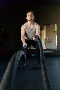 Focused man in gym using battle ropes for strength training, showcasing his muscular build.